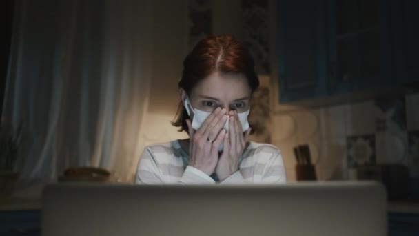 Ung flicka i köket med laptop. Ung kvinna arbetar på distans hemma på grund av en pandemi. Kvinna i medicinsk mask. — Stockvideo