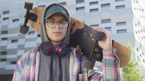 Молодой парень стоит на улице, держа скейтборд на спине. Симпатичный парень в очках и кепке. Позади него многоэтажное здание. — стоковое видео