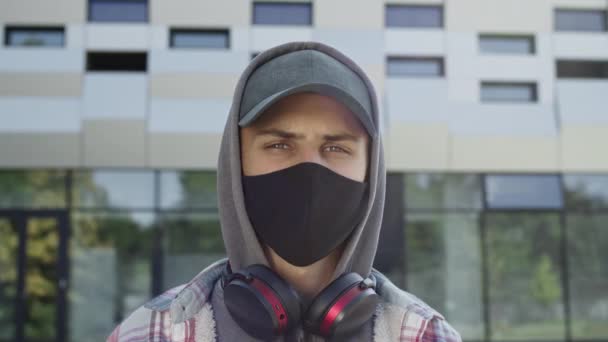 Cara de jovem na máscara preta para se proteger contra vírus. Ele fica na rua de boné e capuz. — Vídeo de Stock