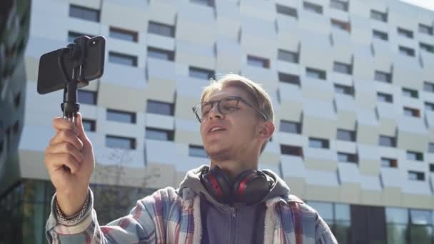 Young Guy Holding Phone And Talking To Someone. He Is Greeted By Waving His Hand. Guy On The Street. Cute Young Guy With Headphones Around His Neck And Wearing Glasses. — Stock Video