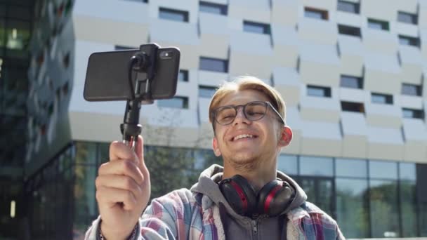 Jongeman die telefoon vasthoudt en met iemand praat. Hij wordt begroet door met zijn hand te zwaaien. Een man op straat. Schattige jonge man met koptelefoon om zijn nek en het dragen van een bril. — Stockvideo