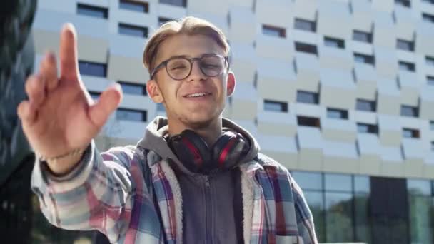 Fröhlicher junger Kerl im Straßengespräch. Blond Guy In Brille Und Kopfhörer Um Seine Hals. — Stockvideo