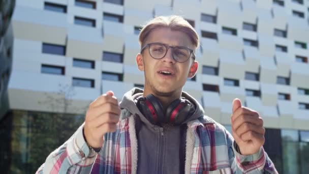 Fröhlicher junger Kerl im Straßengespräch. Blond Guy In Brille Und Kopfhörer Um Seinen Hals. Hinter ihm steht das Bauen. Schöner sonniger Tag. — Stockvideo