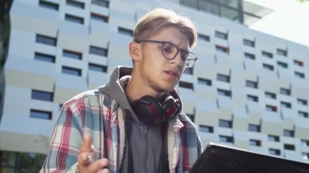 Junger Kerl mit Brille sitzt auf der Straße, Laptop auf dem Schoß und unterhält sich aktiv mit jemandem per Videolink. Er erklärt etwas, indem er mit seiner Hand aktiv gestikuliert. — Stockvideo