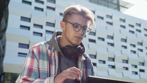 Jovem com óculos sentado na rua com laptop. Ele tira os óculos, limpa os olhos e depois coloca-os de novo.. — Vídeo de Stock