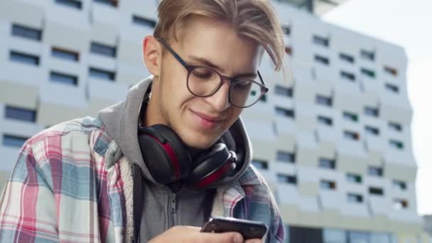 젊은 남자가 거리에서 폰을 들고 있다. 안경을 쓴 귀여운 금발의 남자 - 전화기를 사용하다. — 비디오