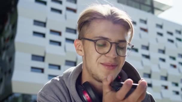 Młody facet z telefonem na ulicy. Słodki blondyn w okularach używa telefonu. Uśmiecha się.. — Wideo stockowe