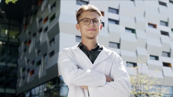 Man in witte jas staat op de achtergrond van Tall Building. Jonge man met een bril. Hij is wetenschapper of arts.. — Stockvideo