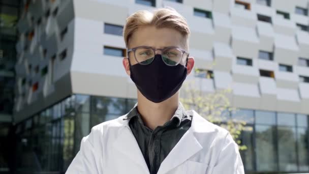Facet w białym płaszczu stoi na tle wysokiego budynku. Młody facet w okularach i w masce do ochrony przed wirusami. Jest naukowcem albo lekarzem.. — Wideo stockowe