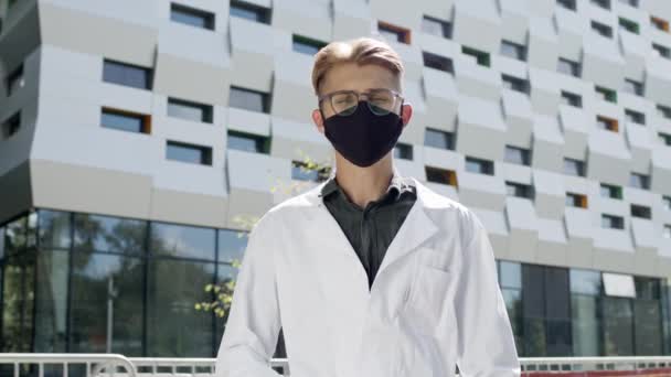 Mann im weißen Mantel steht auf dem Hintergrund eines hohen Gebäudes. Junger Mann mit Brille. Er ist Wissenschaftler oder Arzt. — Stockvideo