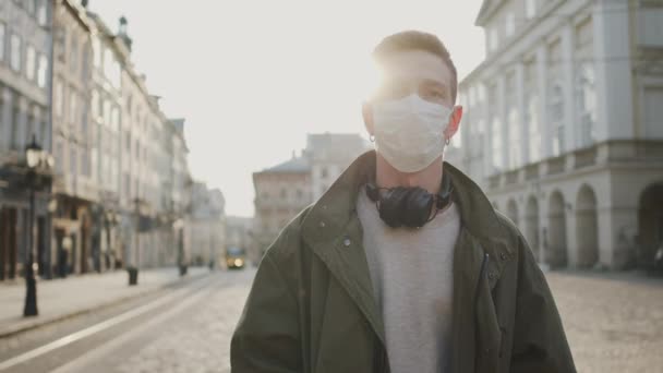 Ο νεαρός με την ιατρική μάσκα που περπατάει στο δρόμο. Η οδός φρουρείται από πανδημία. Ο τύπος φοράει ακουστικά.. — Αρχείο Βίντεο