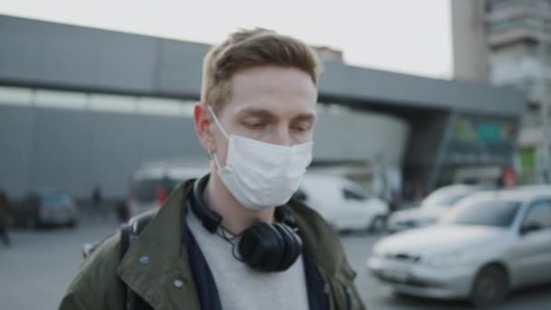 Fiatal fickó orvosi maszkban sétál az utcán. Az utcát a járvány miatt őrzik. A fickó fülhallgatót visel.. — Stock videók