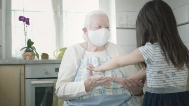 Grootmoeder knuffelt haar kleindochter zittend in de keuken. Ze dragen allebei een medisch masker.. — Stockvideo