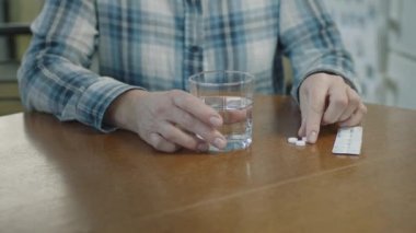 Kadın, mutfakta otururken hap yutar ve su içer..