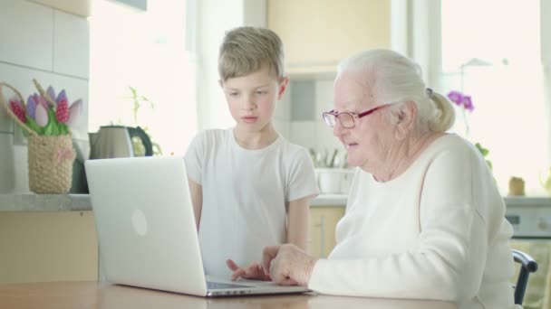 Avó de cabelos grisalhos em óculos olha cuidadosamente para a tela do laptop. Ela é ajudada pelo pequeno bisneto. — Vídeo de Stock