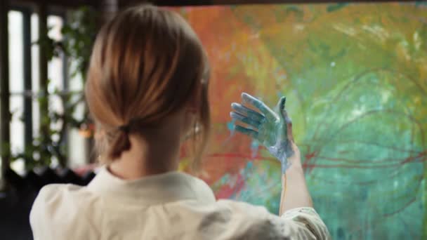 Chica artista en el estudio de arte. Ella mira sus manos empapadas en pintura. Vista desde la parte posterior. — Vídeos de Stock