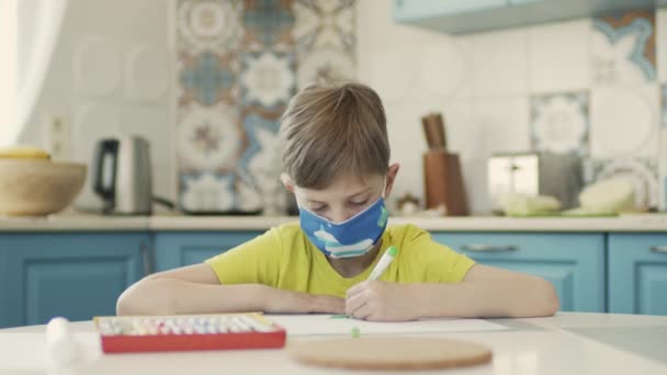 Chlapec v lékařské masce sedí u stolu v kuchyni a kreslí. — Stock video