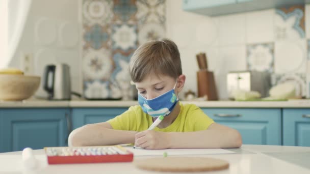 Niño en máscara médica se sienta en la mesa en la cocina y dibuja. — Vídeos de Stock