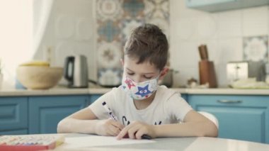 Tıbbi maskeli çocuk mutfaktaki masaya oturur ve resim çizer..
