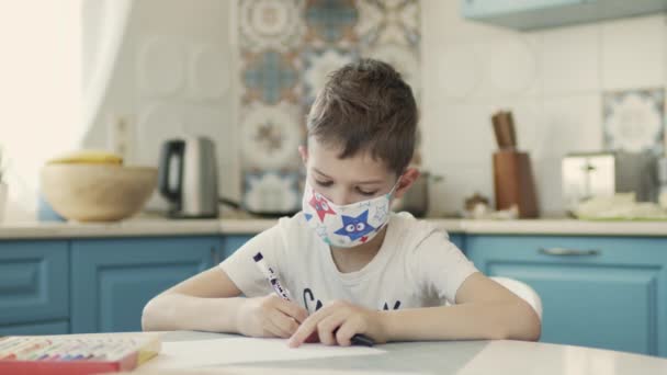 Chlapec v lékařské masce sedí u stolu v kuchyni a kreslí. — Stock video