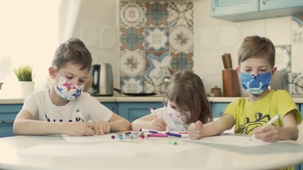 Két fiú és a kishúguk sokszínű orvosi maszkban rajzolnak az asztalra a konyhában.. — Stock videók
