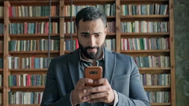 Junger Mann in der Bibliothek spielt emotional Spiel am Telefon. Netter junger Mann aus dem Nahen Osten. Stilvoller Kerl mit Bart im Sakko. — Stockvideo