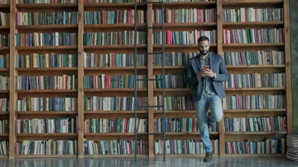 Стильний молодий чоловік в бібліотеці з мобільним телефоном в руках . — стокове відео