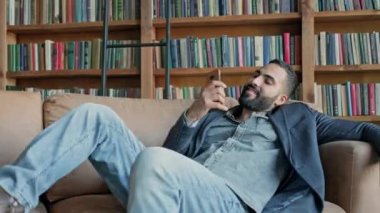 Kütüphanede elinde cep telefonuyla koltukta oturan genç adam..