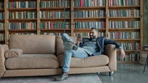 Jeune homme assis sur le canapé dans la bibliothèque avec téléphone portable dans ses mains. — Video