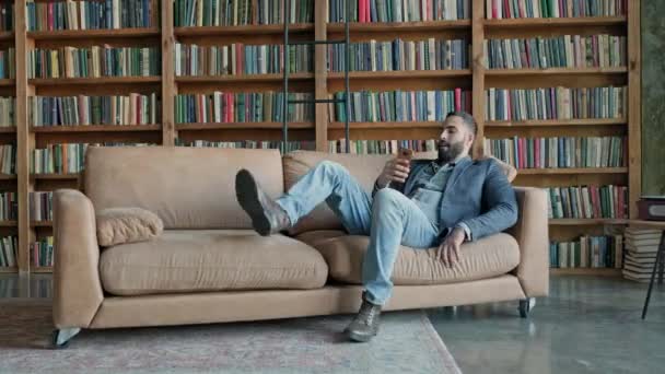 Jeune homme assis sur le canapé dans la bibliothèque avec téléphone portable dans ses mains. — Video