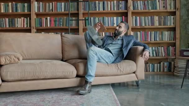 Jeune homme assis sur le canapé dans la bibliothèque avec téléphone portable dans ses mains. — Video