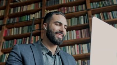 Koyu renk saçlı adam bir sürü kitabı olan bir odada dizüstü bilgisayar kullanıyor..