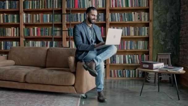 Élégant sombre chevelu gars en utilisant ordinateur portable dans la chambre avec beaucoup de livres. — Video