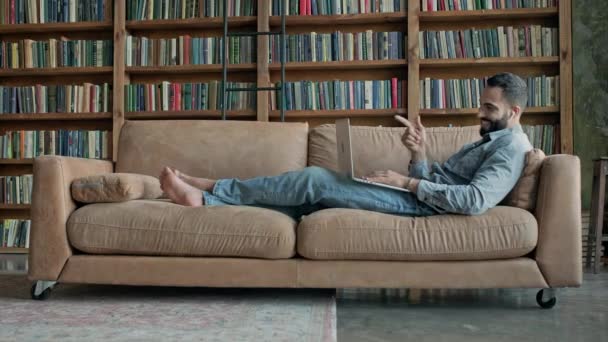 O cara está deitado no sofá e conversando via laptop com alguém. — Vídeo de Stock