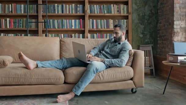 Hombre joven descalzo sentado en el sofá en posición relajada con el ordenador portátil. — Vídeo de stock