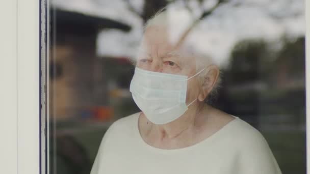 Portret staruszki w masce medycznej. Kobieta wyglądająca przez okno. — Wideo stockowe