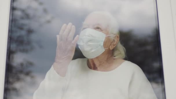 Retrato da velha mulher na máscara médica. Mulher a olhar pela janela. Mulher toca seus lábios com a mão como se estivesse enviando um beijo de ar. — Vídeo de Stock