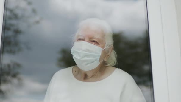 Egy idős nő portréja orvosi maszkban. Egy nő néz ki az ablakon.. — Stock videók