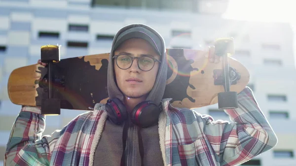 Giovane ragazzo in piedi sulla strada tenendo Skateboard sulla schiena. Un ragazzo carino con occhiali e cappello. Dietro di lui c'è l'edificio a più piani. — Foto Stock