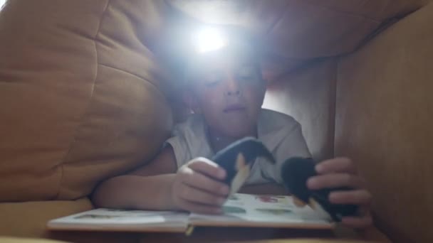 Niño con linterna en la cabeza lee libro. — Vídeos de Stock