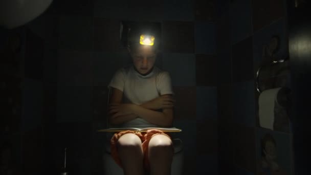 Chłopiec siedzi w toalecie z latarką w ręku i czyta książkę.. — Wideo stockowe