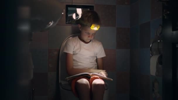 Мальчик сидит в туалете с фонариком в руках и читает книгу. — стоковое видео