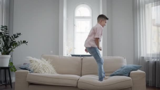 Junge hat Spaß. Er springt auf die Couch mit Kissen in den Händen. — Stockvideo