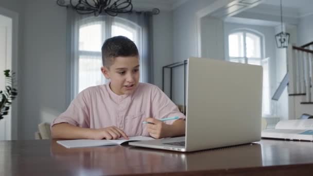 Chłopiec Osiem lat siedzi przed laptopem przy stole. Pisze w notatniku. Chłopiec robi prace domowe. — Wideo stockowe