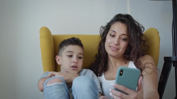 Junge Mutter und Sohn sitzen zusammen im Stuhl. Frau hält Telefon und sie sprechen mit jemandem. — Stockvideo