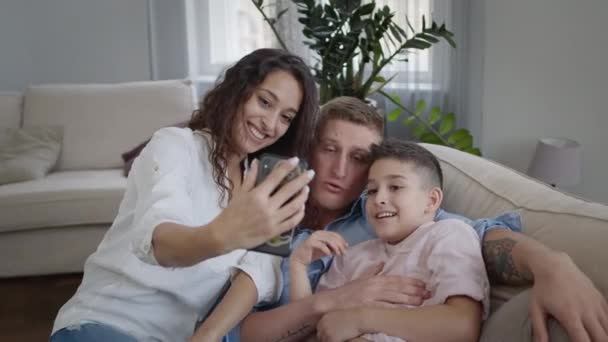 Mladá matka fotí svou rodinu na telefonu. Máma, táta a syn na gauči. Ukazují legrační výrazy obličeje. Rodina baví. — Stock video