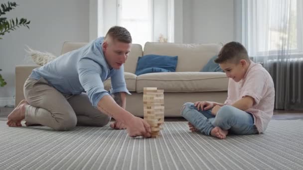 Батько і син сидять на підлозі в приміщенні. Вони грають у збирання піраміди дерев "яних баранів. — стокове відео