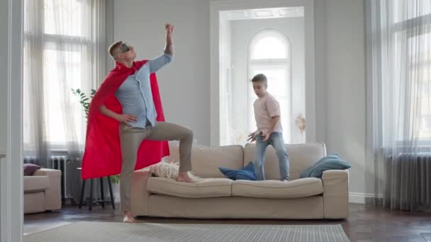 Vader en zoon in het appartement spelen spel. Vader in rode mantel en bril vertegenwoordigt Superman. — Stockvideo
