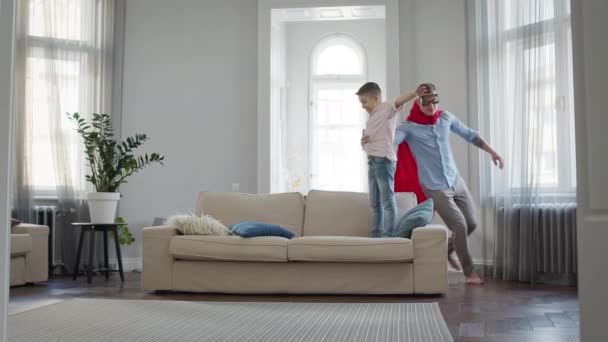 Padre e hijo en el apartamento jugando juego. Papá con capa roja y gafas representa a Superman. — Vídeo de stock