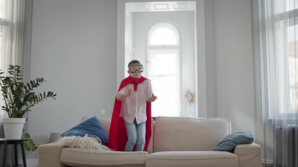 Pojken står på soffan i lägenheten. Han bär röd mantel och glasögon. Han spelar Stålmannen. — Stockvideo